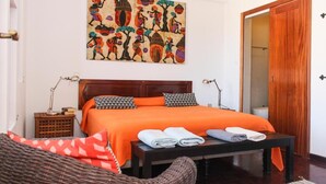 A bedroom with a wooden bed, orange bedding, a wall painting, a wooden door, and a wicker chair. - Vila Mira Mar (São Vicente)