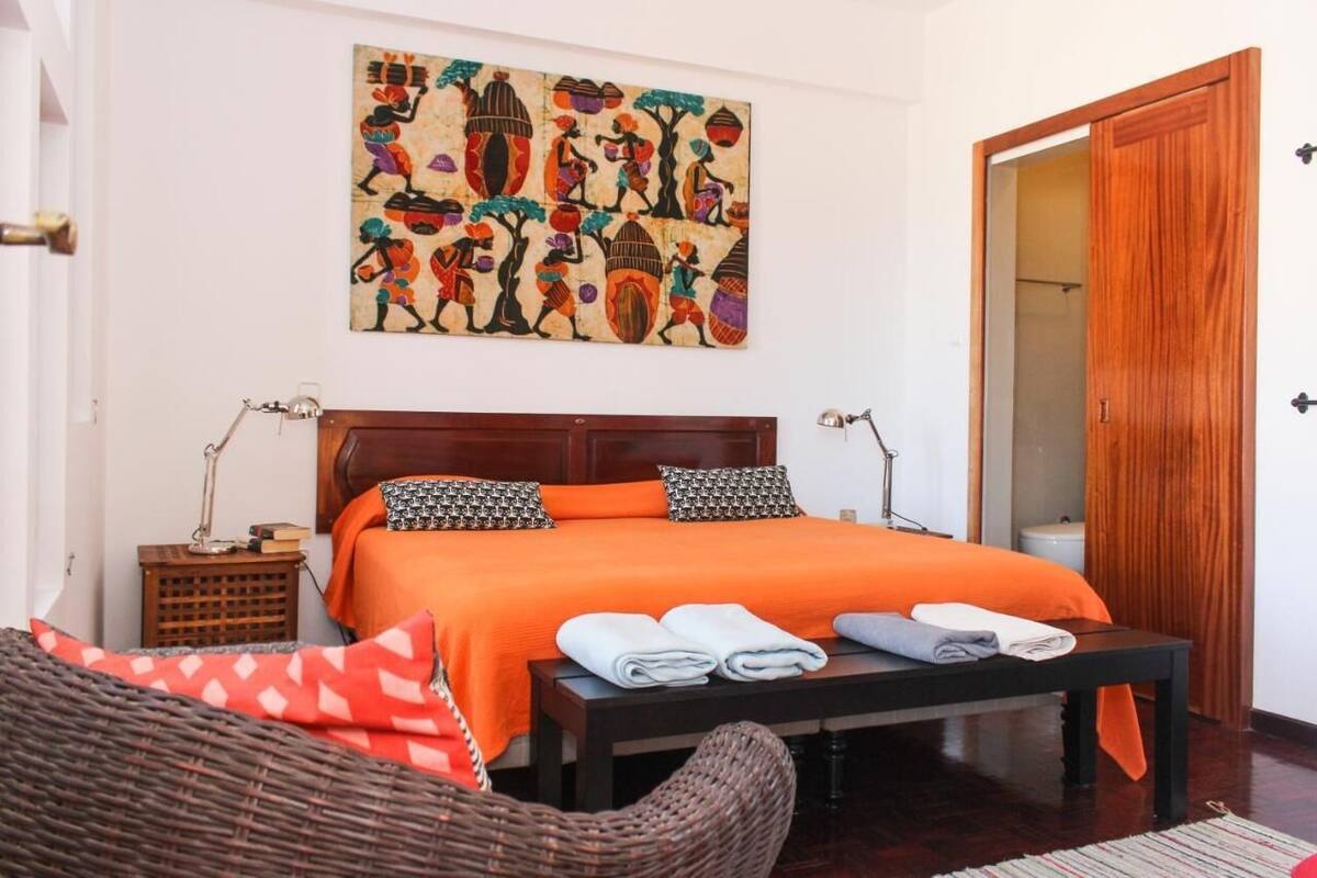A bedroom with a wooden bed, orange bedding, a wall painting, a wooden door, and a wicker chair.