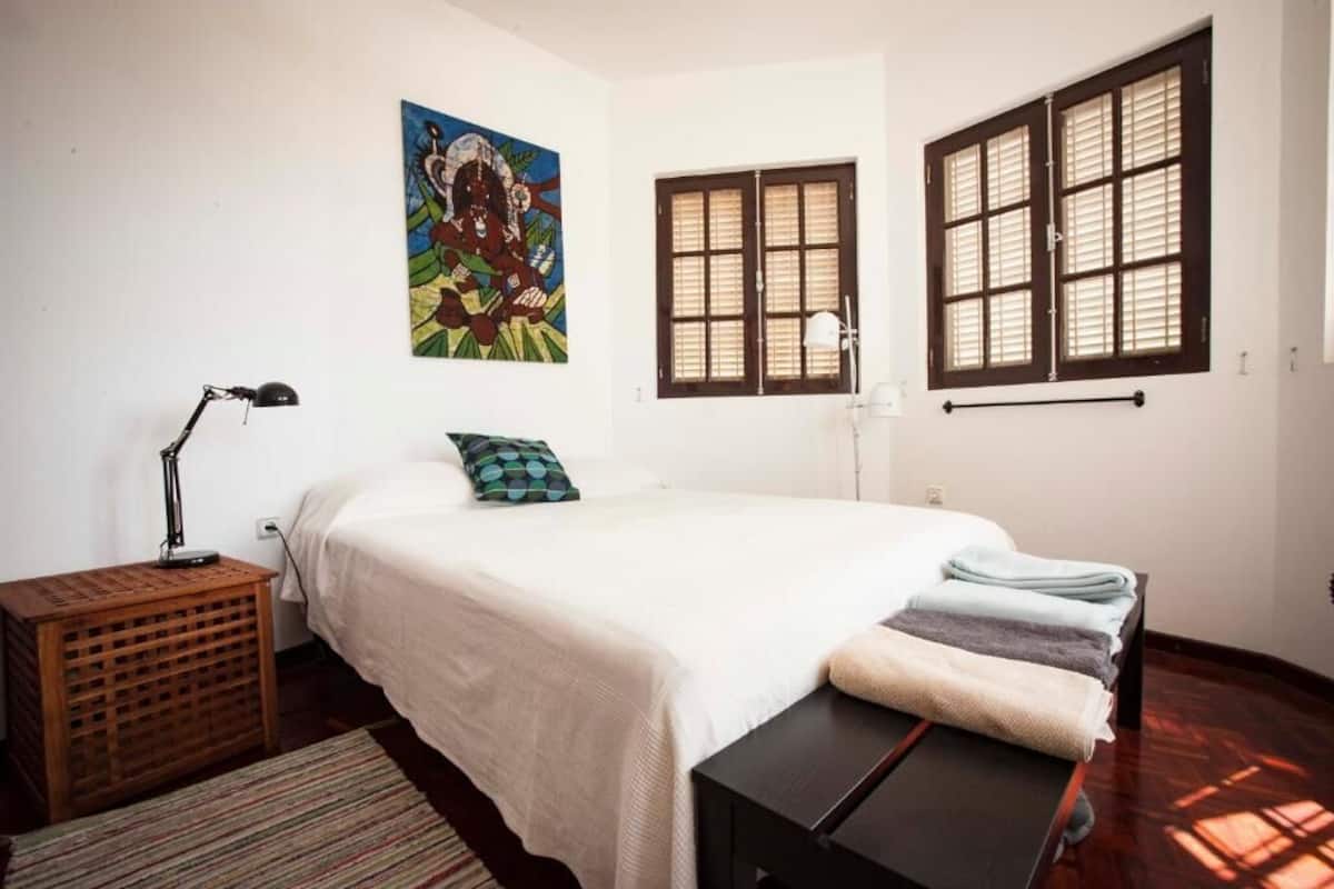 A bedroom with a bed, a wooden side table, a lamp, a painting, and two windows with shutters.