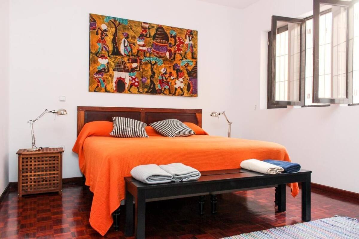 A bedroom with a large bed, a wooden headboard, a colorful abstract painting, a window with blinds, and a small table with towels.