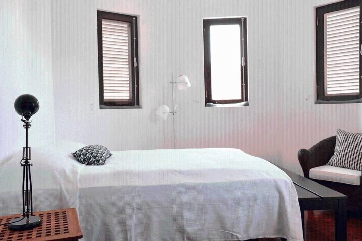 A hotel room with a bed, two windows with blinds, a desk, a chair, and a lamp.