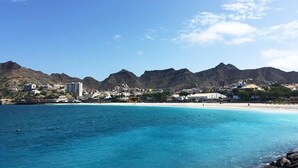 A coastal town with a beach, clear blue waters, and mountains in the background. - Vila Mira Mar (São Vicente)
