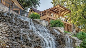 Une cascade d’eau dévalant une pente rocheuse, se jetant dans une étendue d’eau au pied d’une terrasse en bois surmontée d’une pergola.
