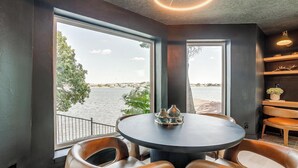 Un espace de repas moderne avec une table ronde et des chaises en cuir, de grandes fenêtres offrant une vue sur un plan d’eau, et une étagère ornée d’objets décoratifs.