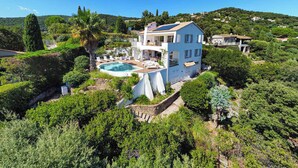Une grande maison avec une piscine, entourée d’une végétation luxuriante et de collines.