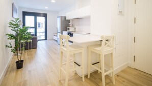 Una cocina moderna con armarios blancos, un refrigerador y un microondas. Hay un comedor con una mesa blanca y dos sillas. Se ve una planta en maceta en el suelo.