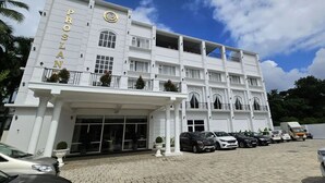 A multi-story building with a white facade and the name "PROSLAN" prominently displayed. There are multiple balconies with railings and potted plants. The entrance features a covered area with a glass door. Several cars are parked in front of the building.