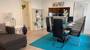 A modern living room with a grey sofa, a dining table with black chairs, a blue rug, and a kitchen area with wooden cabinets and a wine rack.