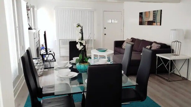 A modern living room with a glass dining table, black chairs, a sofa, and a small side table with a lamp.