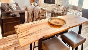 Una sala de estar con chimenea, ventilador de techo, televisor montado arriba y una mesa de comedor con taburetes.