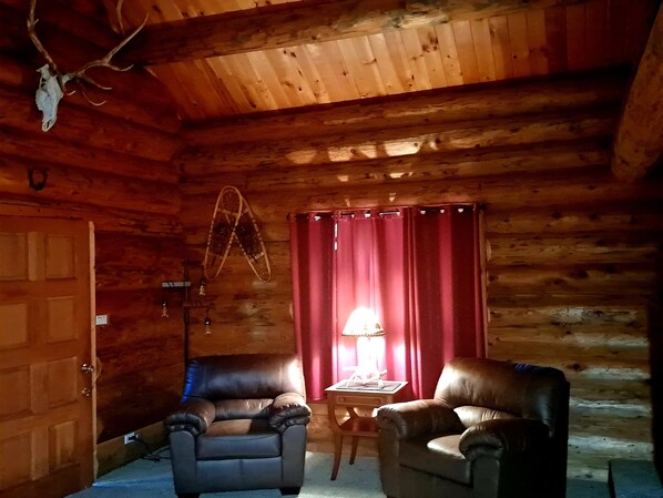 L'intérieur d'un chalet en bois, avec deux fauteuils en cuir, une petite table avec une lampe et un rideau rouge.