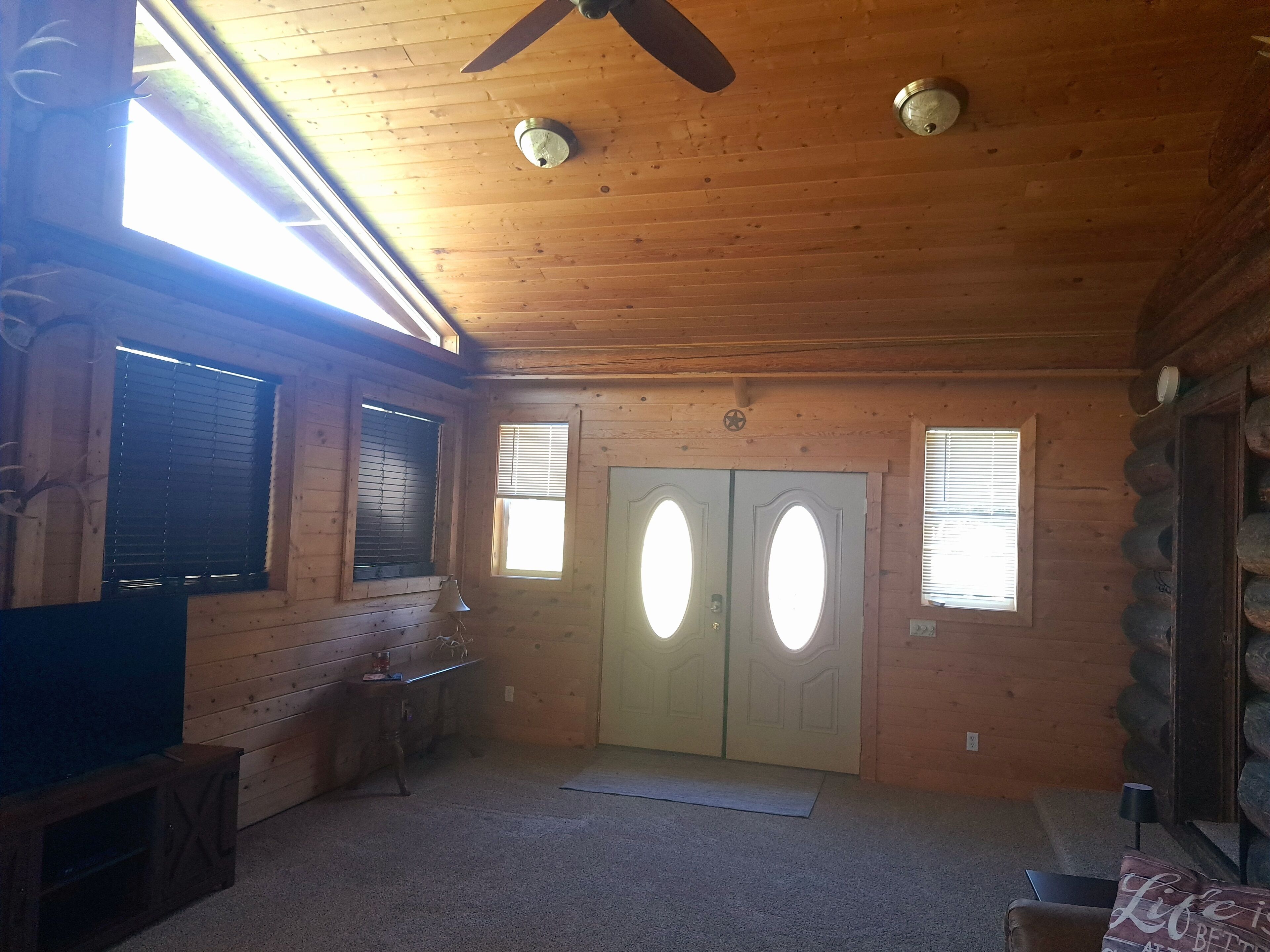 Interno di una baita con soffitto in legno, due porte con finestre ovali, una ventola a soffitto e un tavolino con una lampada.