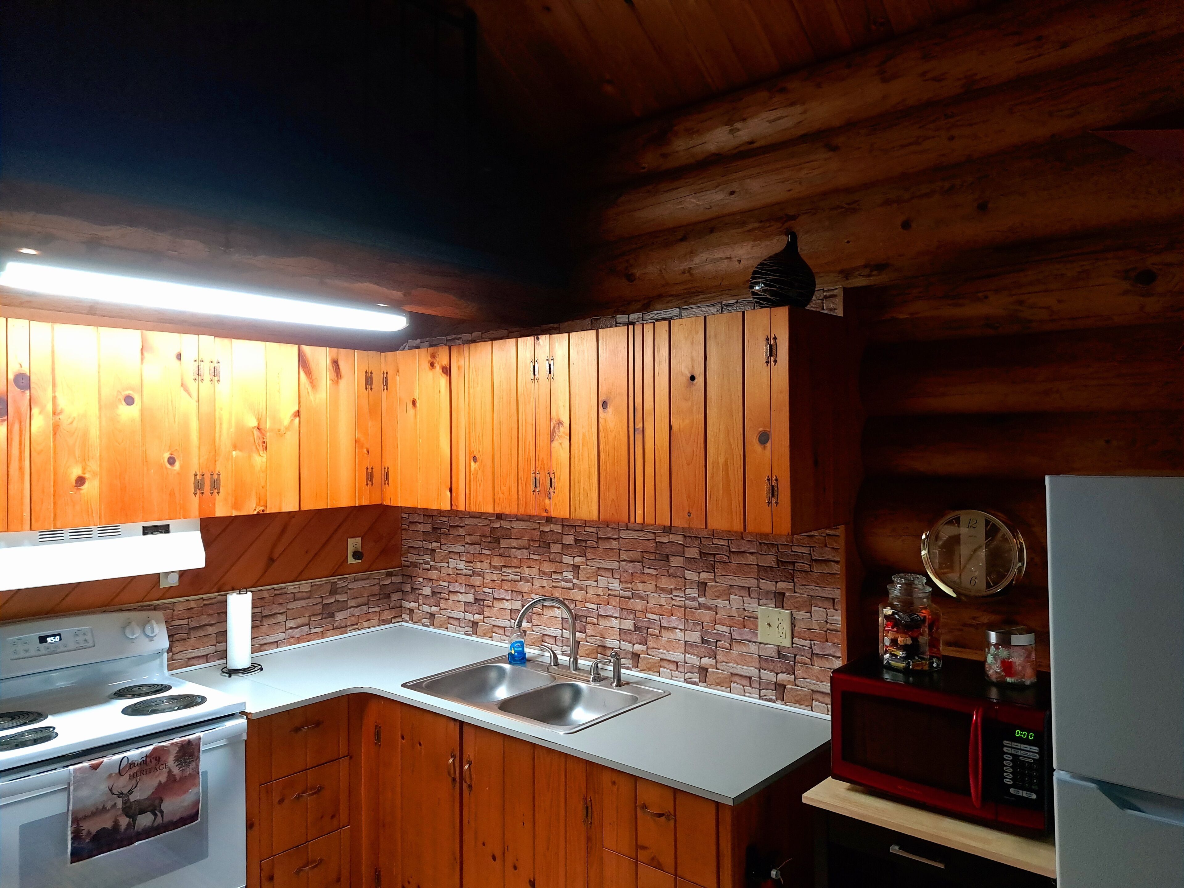 A kitchen with wooden cabinets, a white countertop, a stove, a sink, a microwave, and a clock on the wall.