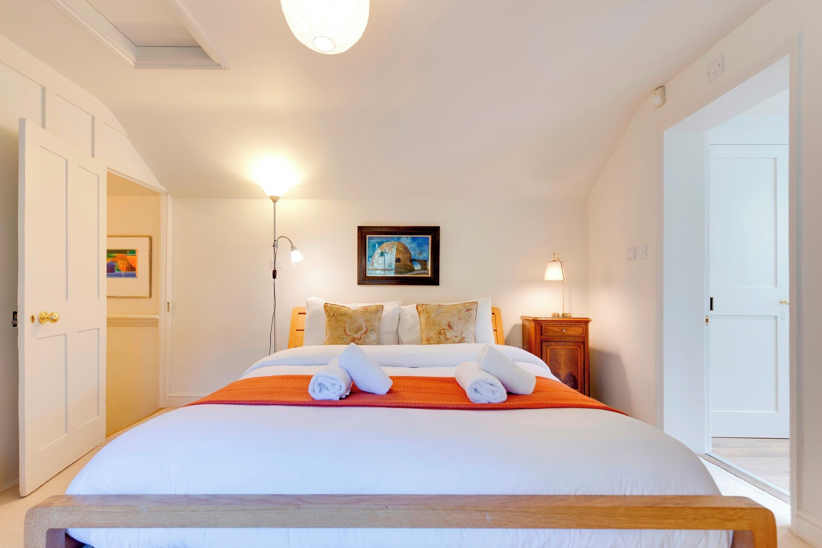 A neatly arranged bedroom with a large bed, two bedside lamps, a framed picture on the wall, and a door leading to another room.