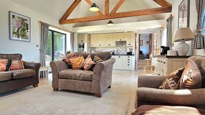 A spacious living area with a high ceiling and exposed beams, a cozy sofa set, a kitchen in the background, and a view of the outdoors.