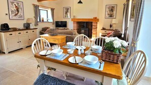 A cozy living room with a fireplace, wooden beams, a dining table set for four, and a television in the background.
