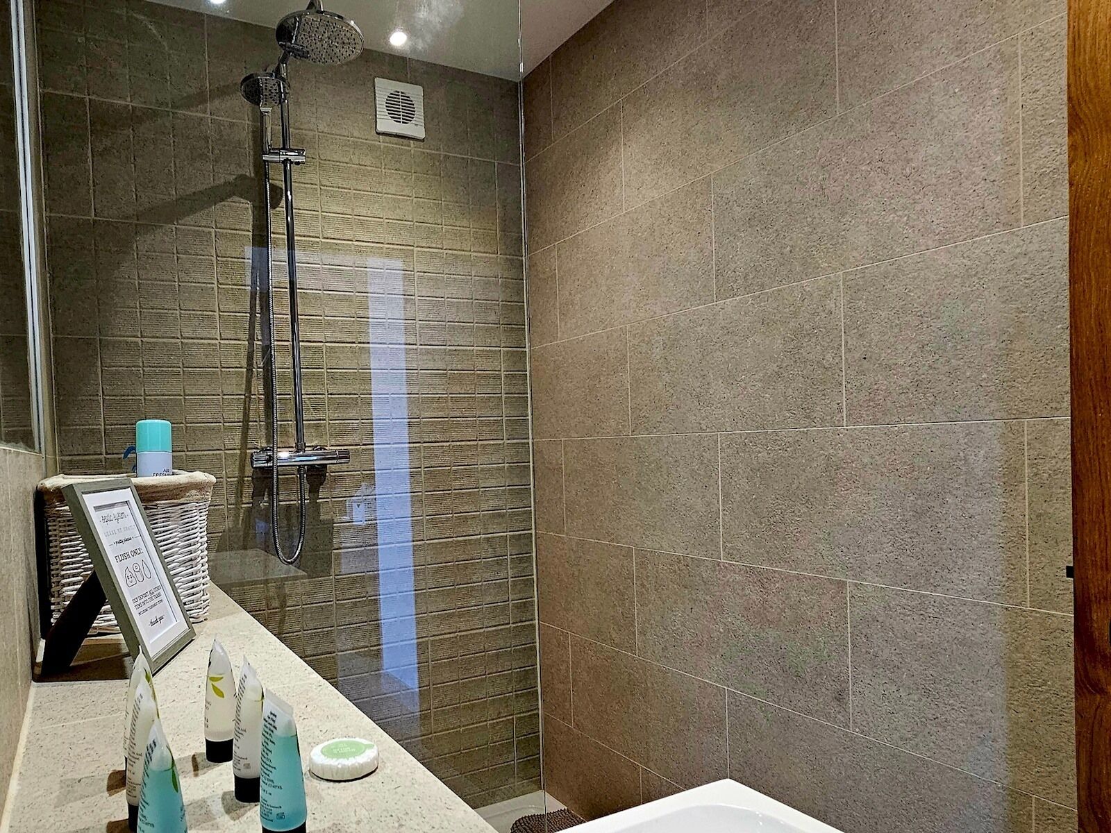 A modern bathroom with a shower, a sink with toiletries, and a framed sign.