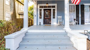 A porch with steps leading to a door, a hanging American flag, and a white picket fence. - Central Location Historic Grandeur (Mobile)