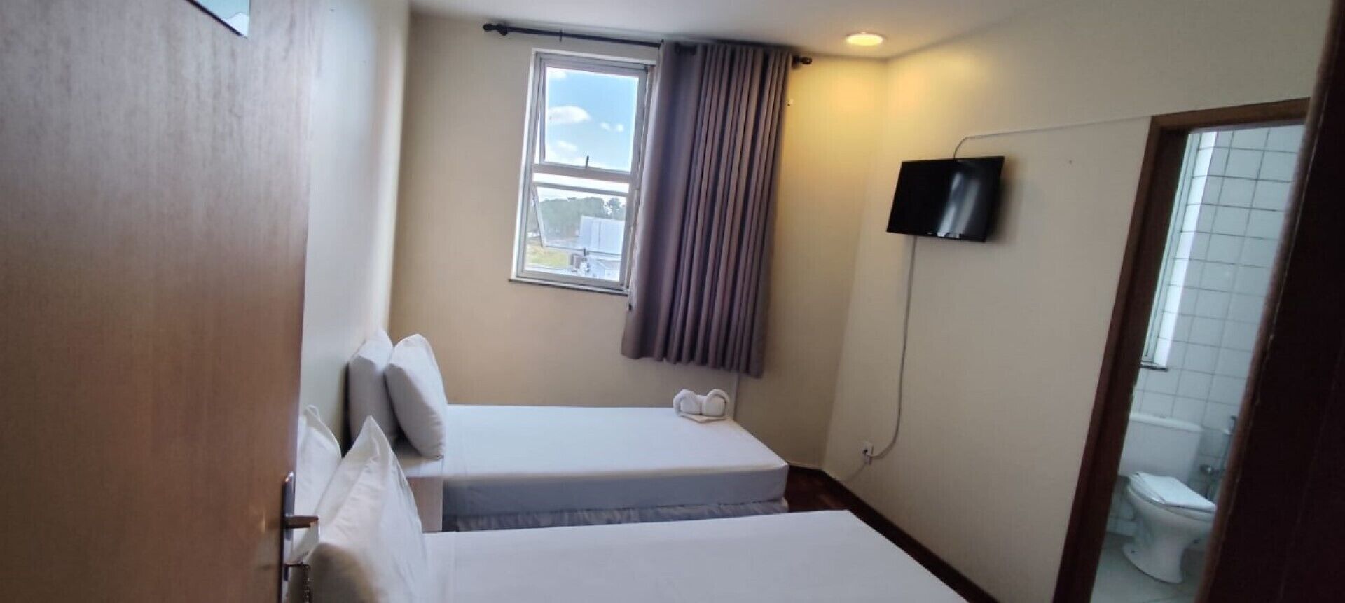 Ein kleines Hotelzimmer mit zwei Betten, einem Fenster mit Vorhängen, einem an der Wand befestigten Fernseher und einer sichtbaren Toilette durch eine offene Tür.