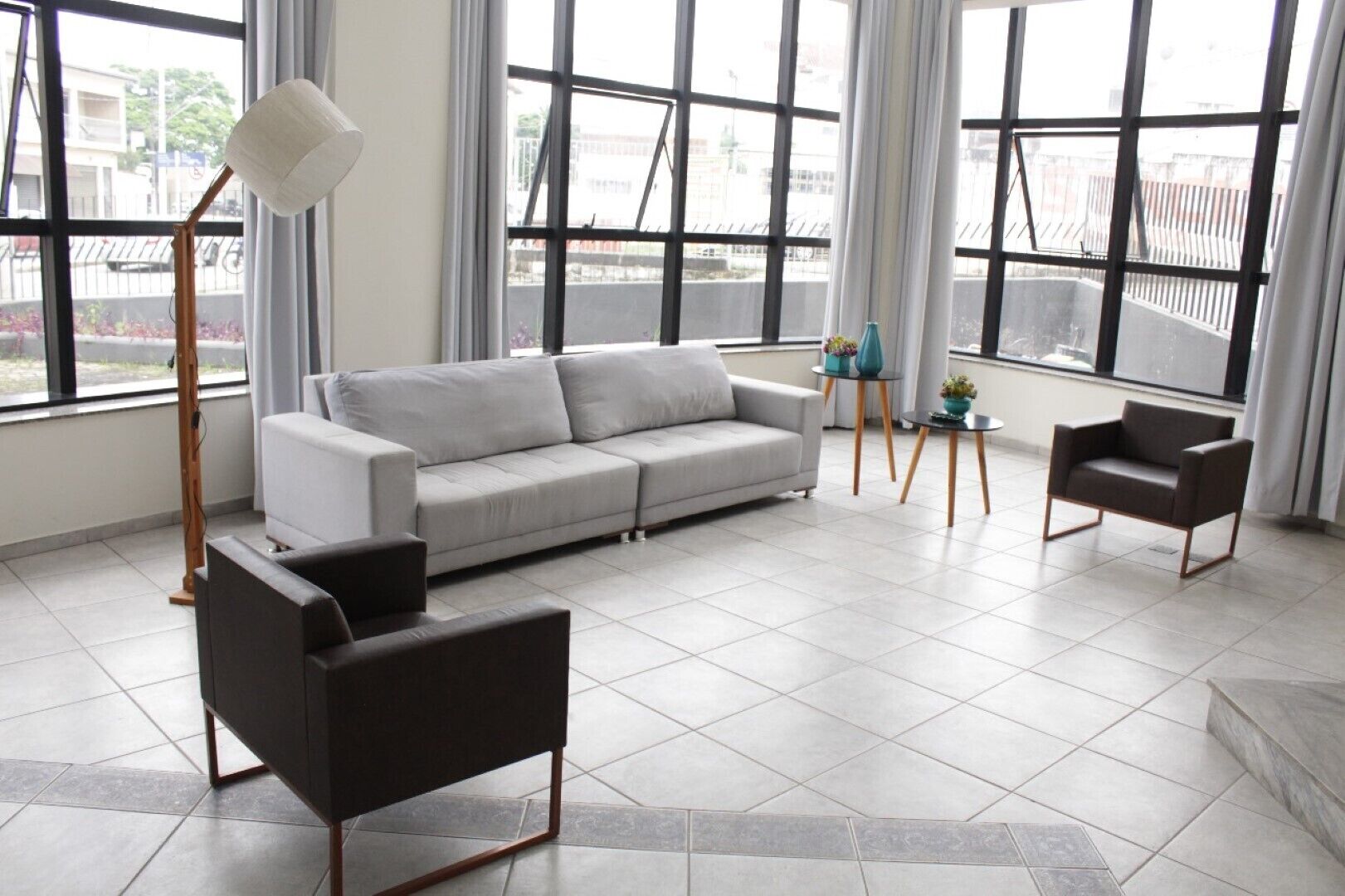A modern living room with a grey sofa, a black armchair, a floor lamp, and a small table with decorative items. Large windows provide a view of the outside.