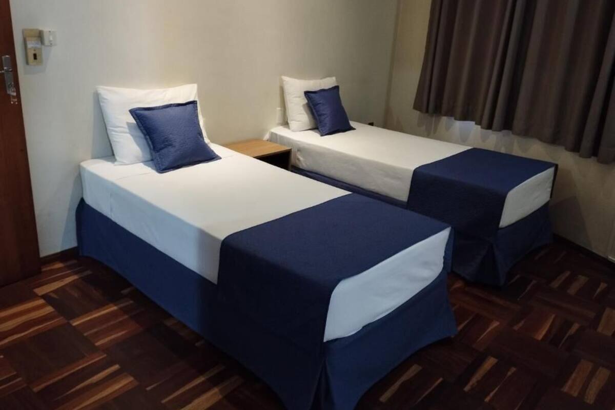 Dos camas individuales con ropa de cama blanca y azul, piso de madera y una ventana con cortinas.