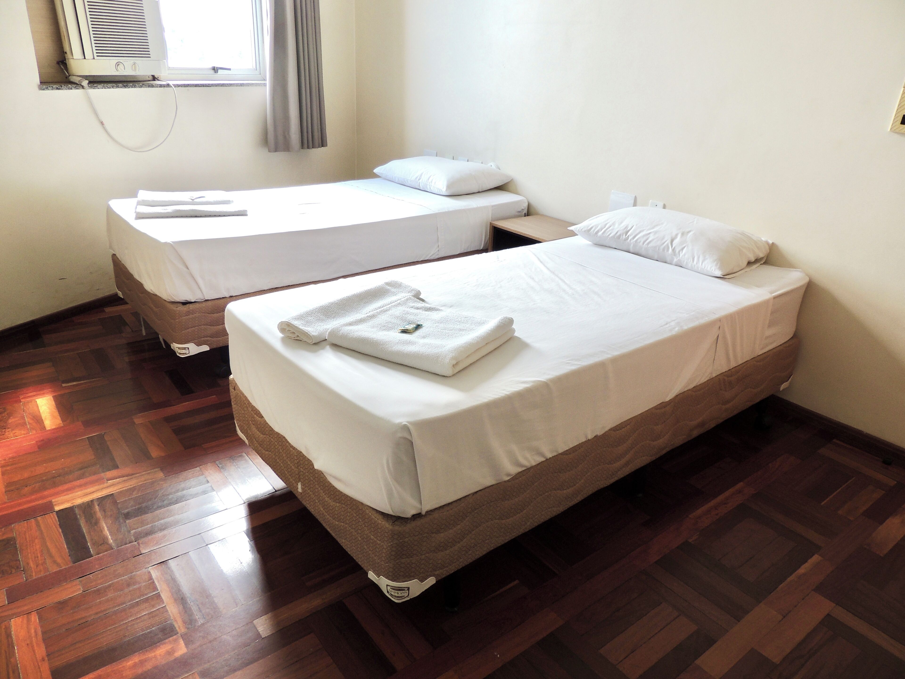 Two single beds with white linens and pillows, a wooden parquet floor, and a window with a curtain.