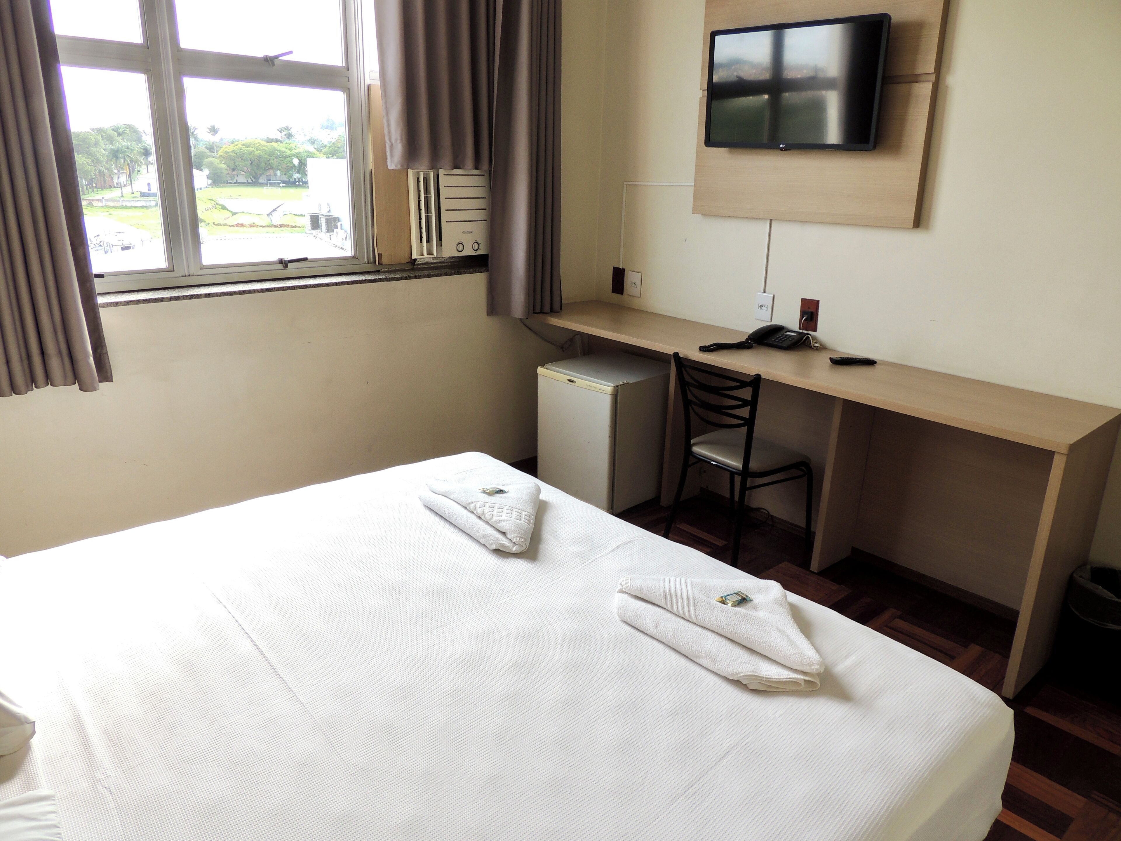 Une chambre d’hôtel avec un lit, un bureau, une chaise, une télévision et une fenêtre avec des rideaux.