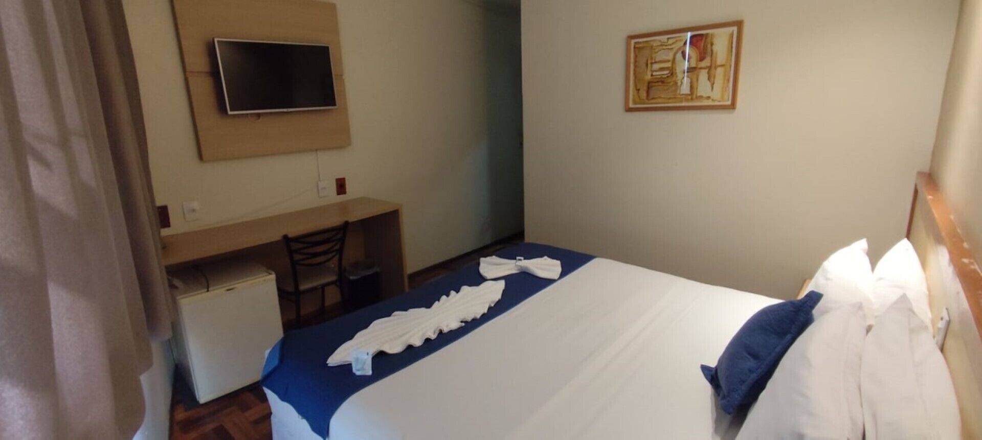 A hotel room with a bed, a desk, a chair, a flat-screen TV mounted on the wall, and a painting on the wall.