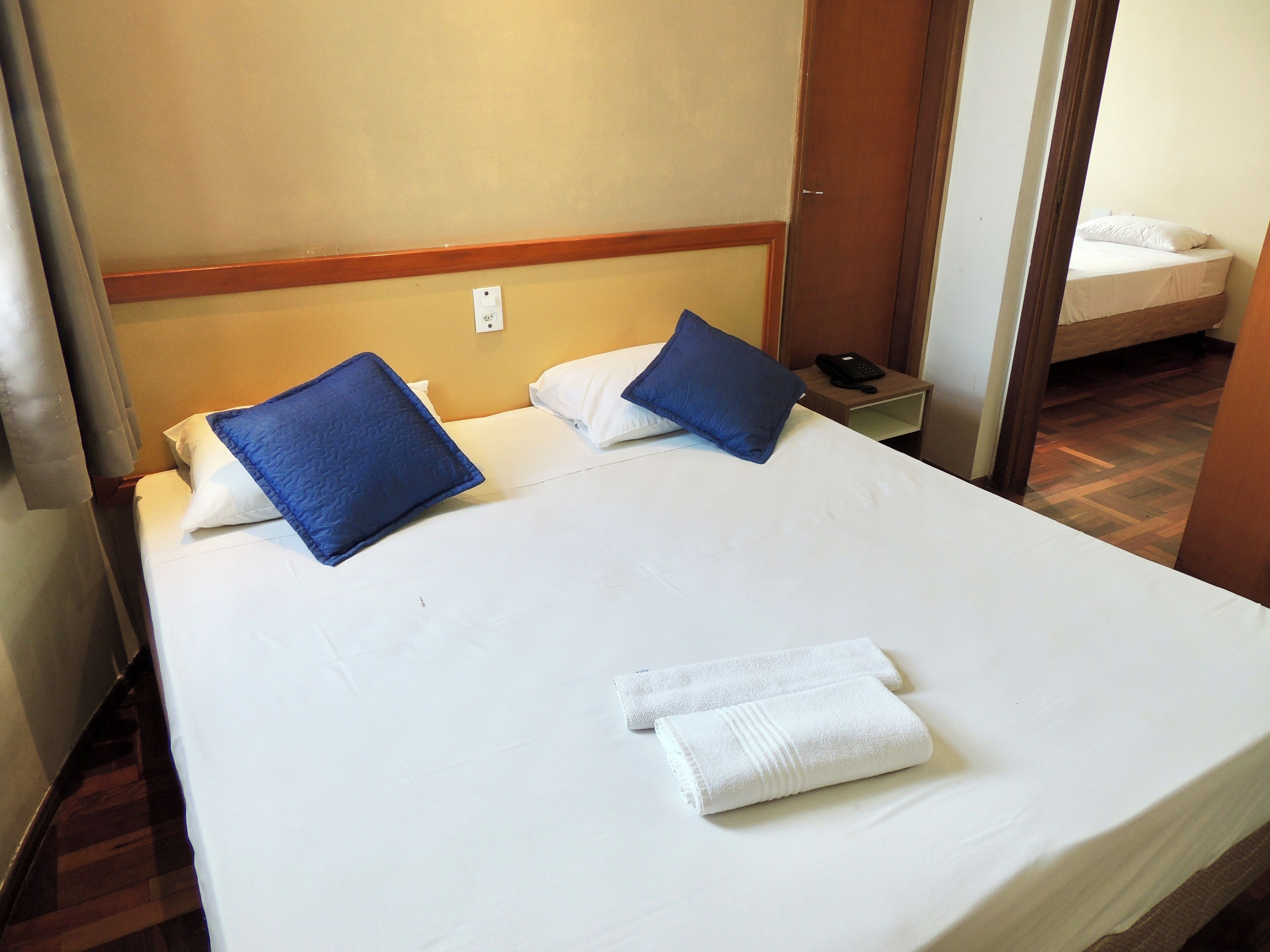Una cama doble con ropa de cama blanca y azul, una mesita de noche con un teléfono, y suelo de madera.