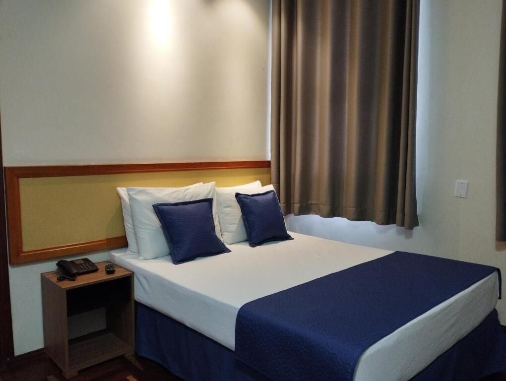 Een hotelkamer met een bed, nachtkastje, telefoon en een raam met gordijnen.