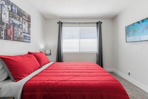 Een slaapkamer met een rood beddegoed, een schilderij aan de muur, een raam met gordijnen en een televisie.