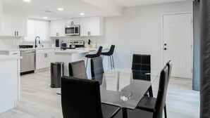 A modern kitchen with white cabinets, a microwave, and a coffee machine. There is a dining area with a glass table and black chairs.
