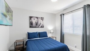 A bedroom with a bed, a nightstand, a lamp, a window with curtains, and artwork on the wall.