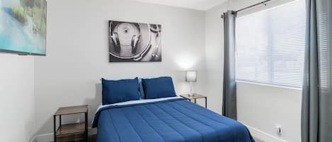Un dormitorio con cama, mesita de noche, lámpara, ventana con cortinas y una obra de arte en la pared.