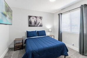 Un dormitorio con cama, mesita de noche, lámpara, ventana con cortinas y una obra de arte en la pared.
