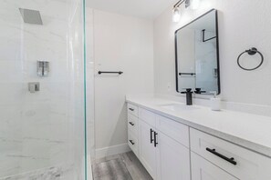 A modern bathroom with a glass shower enclosure, white marble tiles, a large mirror, and a white countertop with black handles.