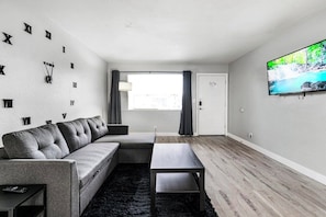 A modern living room with a sectional sofa, a coffee table, a flat-screen TV, and a large window with curtains.