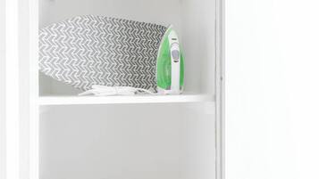 A white shelf with a patterned fabric, a green and white iron, folded towels, and a gray laundry basket.