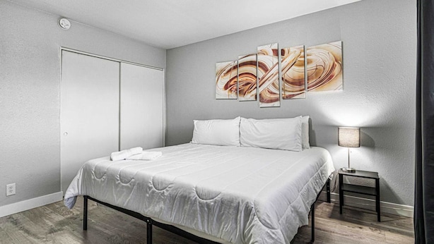 A modern bedroom with a large bed, a bedside table with a lamp, and a wall-mounted abstract painting. - Cozy 1 Bed, Sleeps 4! Near Strip! Perfect GetAway! (Las Vegas)