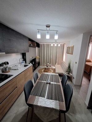 Een moderne keuken met donkere kastjes, een wit aanrechtblad en een eethoek met een tafelkleed met een patroon.