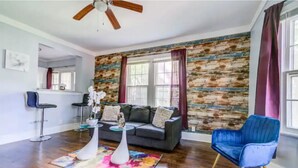 A living room with a wooden wall, a gray sofa, a blue chair, and a ceiling fan. - Cozy King bed and Queen Sofa bed Apt (Decatur)