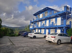 Un immeuble à plusieurs étages aux couleurs bleu et blanc, un espace de stationnement avec plusieurs voitures, et un paysage montagneux en arrière-plan.