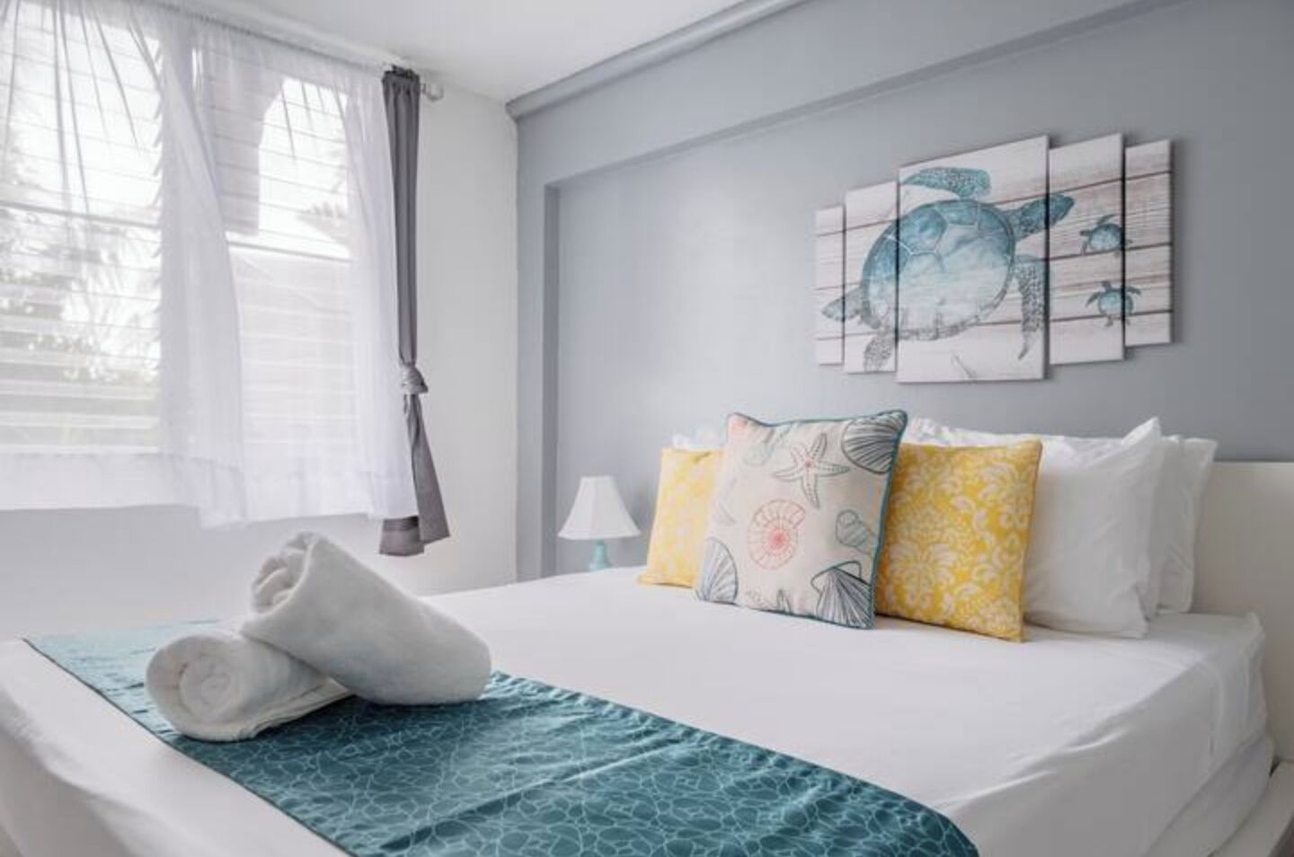 A neatly made bed with a blue patterned bedspread, a pair of rolled towels, and multiple pillows in yellow and white.