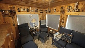 A cozy cabin interior with wooden walls, a ceiling fan, a sofa, two armchairs, a small table with a lamp, and a decorative wall with nautical items. - Lakeside Haven  (Houghton Lake)