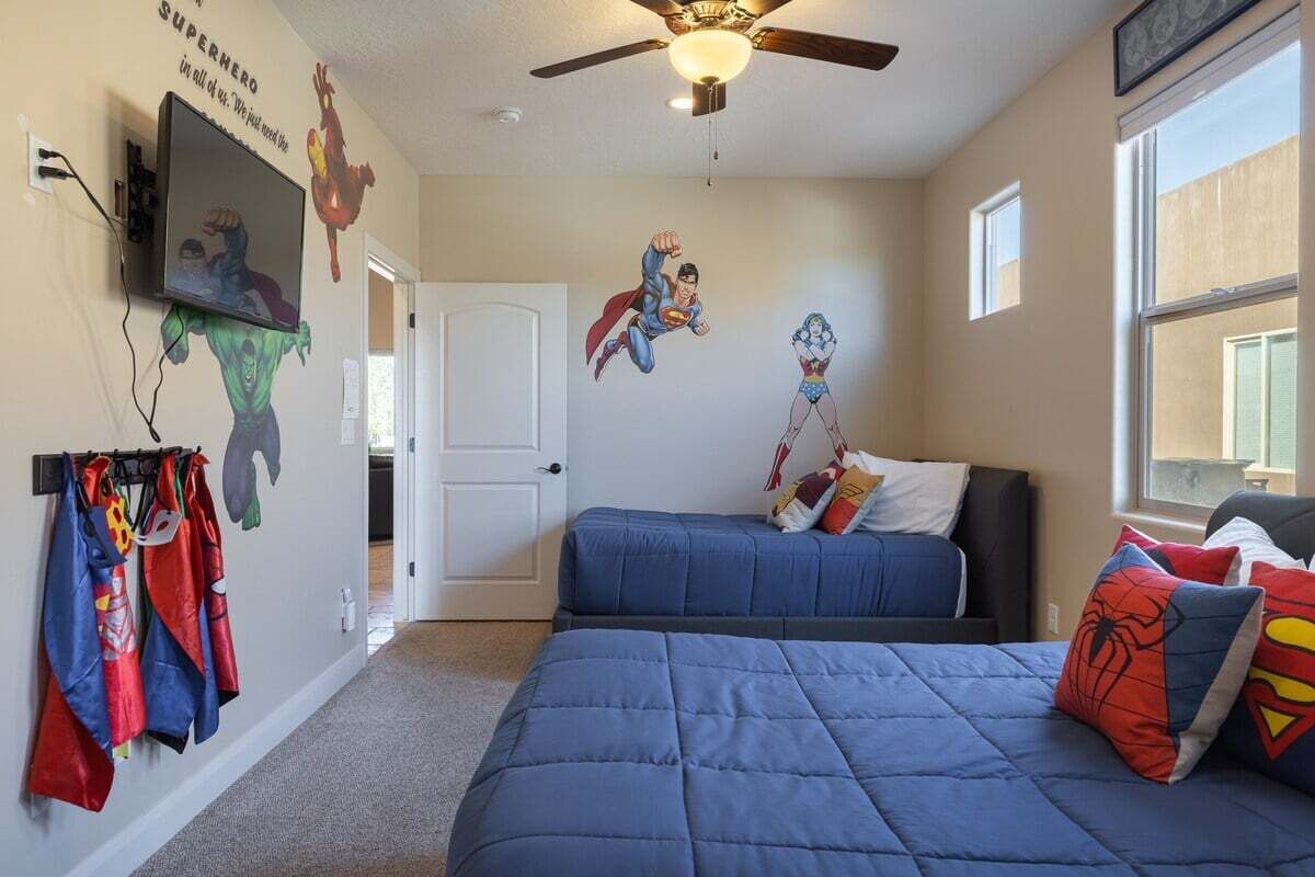 Une chambre avec deux lits, des décorations murales sur le thème des super-héros, un ventilateur de plafond et un téléviseur fixé au mur.