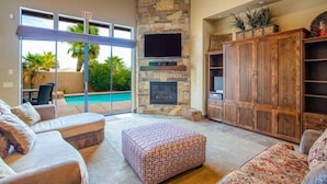 Amplia sala de estar con chimenea de piedra, televisor de pantalla plana y piscina visible a través de puertas de cristal.