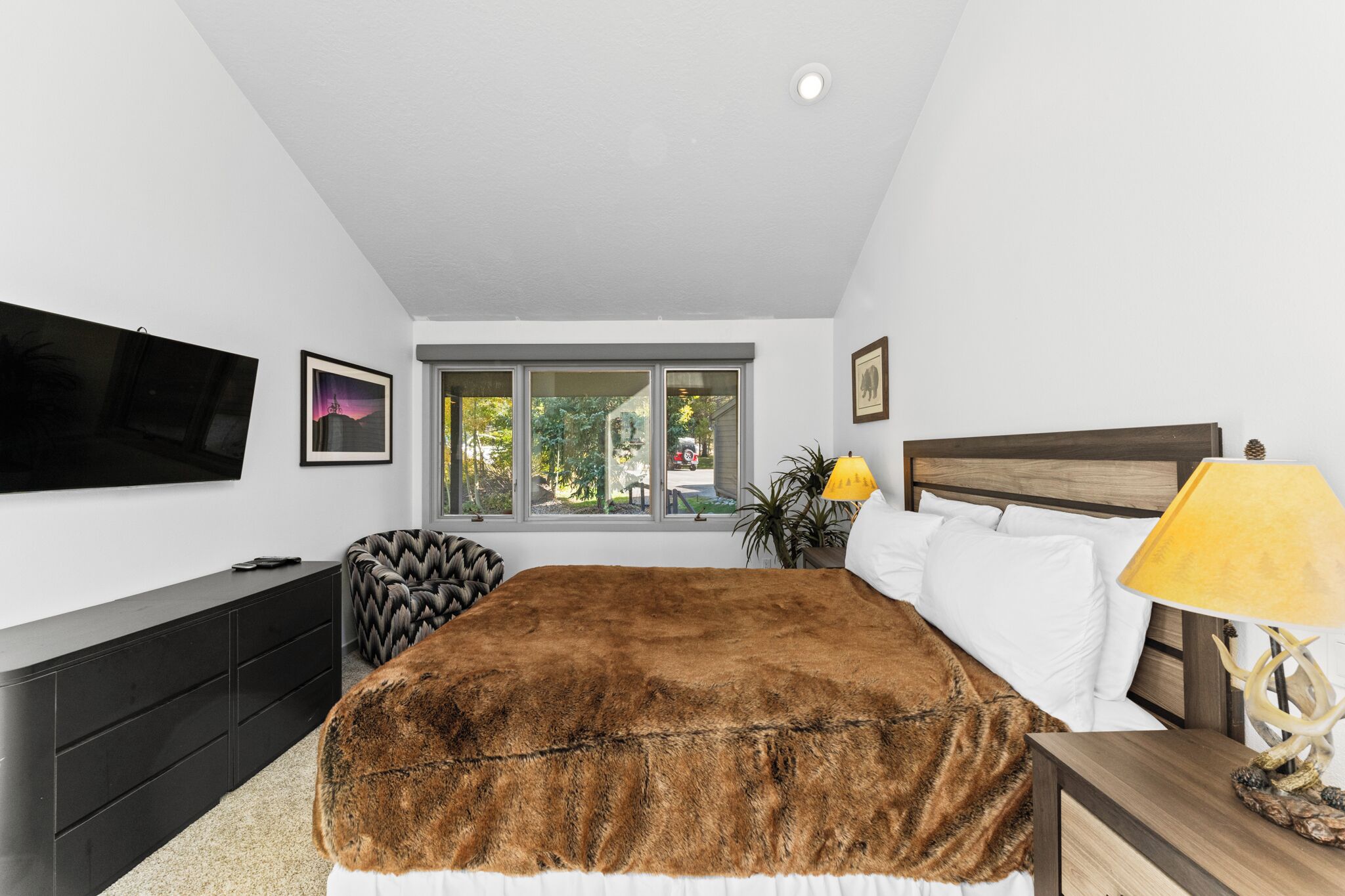 A bedroom with a large bed, a TV mounted on the wall, a window with a view of the outdoors, a bedside table with a lamp, and a black entertainment unit.