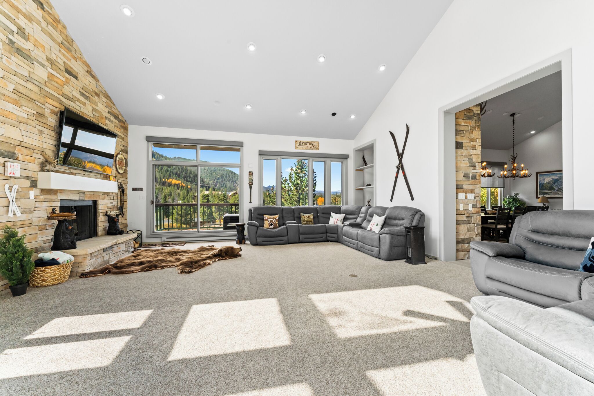 A spacious living room with a stone fireplace, large windows, a grey sectional sofa, and a chandelier.