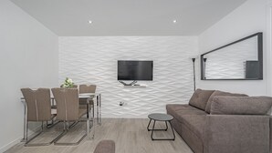 Una sala de estar moderna con un televisor de pantalla plana montado en una pared texturizada, un sofá gris, una mesa de comedor con sillas y una mesita auxiliar.