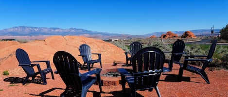 A group of Adirondack chairs arranged on a gravel surface with a view of red rock formations and distant mountains.